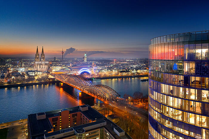 Luftbild der nächtlichen Kölner Innenstadt, auf dem die Hohenzollernbrücke, der Rhein und der Dom zu sehen sind.