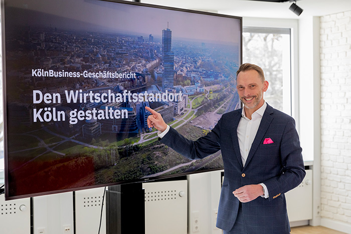 Der Geschäftsführer der KölnBusiness Wirtschaftsförderung steht vor einem Monitor mit der Aufschrift "Den Wirtschaftsstandort Köln gestalten"